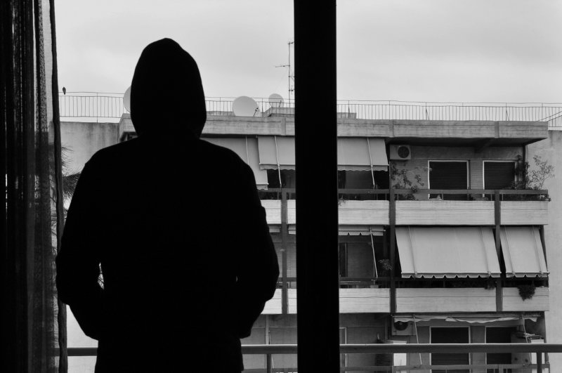 Black and white photo of a hooded man looking out an apartment window